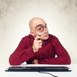 Bald yopung adult man sitting at the table with keyboard holding magnifying glass in front of the computer, internet search concept