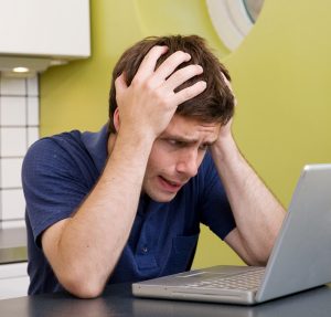 A young male at home in the kitchen looking worried while using a computer.